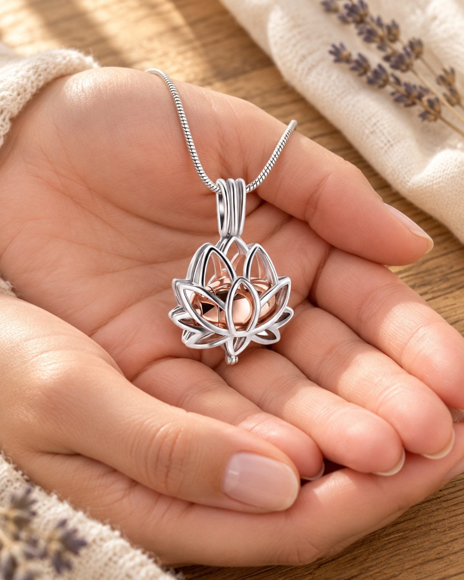 Zwei Hände halten einen Lotus-Erinnerungsanhänger mit roségoldener Innenkammer, daneben elfenbeinfarbenes Leinen und Lavendel